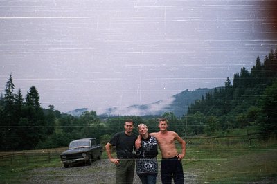 Vintage candid shot of three individuals posing outdoors beside a classic car in a rural, forested setting. The man on the le...