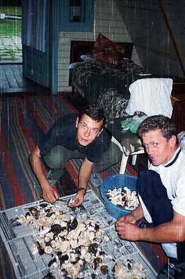 Two men in a dimly lit room prepare seafood, likely mussels, on a metal tray. One man wears a dark shirt, the other a white s...