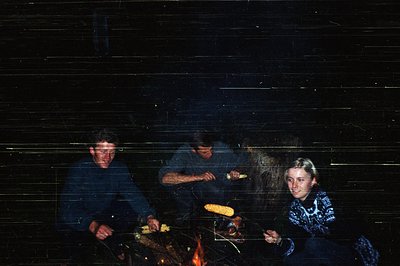 Three individuals roast corn over an open flame in a dimly lit, rustic indoor setting. The wooden walls and rough-hewn beams ...