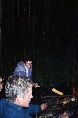 Nighttime outdoor scene featuring two individuals roasting corn on an open flame. One person wears a blue jacket, the other a...