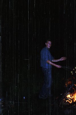 Man stands under cascading water in dimly lit indoor setting, holding a rope. Firelight illuminates right side, suggesting a ...