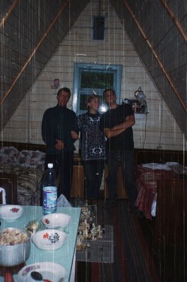 Three individuals pose indoors in a rustic, triangular wooden structure with tiled walls. The central figure wears a patterne...