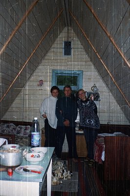 Three individuals pose indoors in a triangular, tiled kitchen with a sloped ceiling. The central figure wears a dark jacket, ...