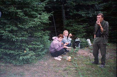 Three individuals in a wooded area, mid-1970s–1980s. Two women kneeling near a small fire pit with a metal pot, one holding a...