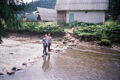 Two individuals stand on a shallow, rocky stream in a rural setting, surrounded by lush greenery and wooden structures. The s...