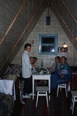 Indoor scene featuring a triangular, tiled loft space with four adults seated around a small white table. The man standing we...
