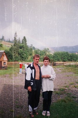 Vintage candid shot of two individuals posing outdoors in a rural, forested setting. The man wears a striped sweater and back...