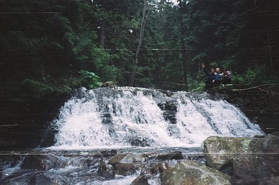 Two individuals rappelling down a cascading waterfall surrounded by dense forest. The scene captures the thrill of outdoor ad...