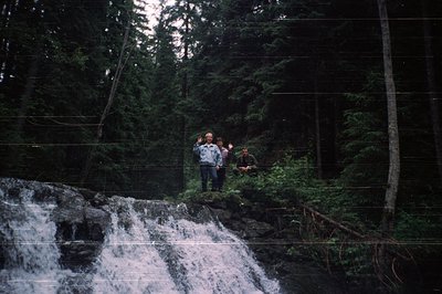 Three hikers pose on rocky ledge beside cascading waterfall in dense conifer forest. Vintage filter enhances moody, sepia-ton...