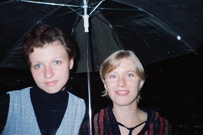 Two women pose under a transparent umbrella in low-light, likely indoors. The woman on the left wears a light gray vest over ...