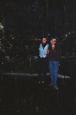 Two individuals pose in front of a reflective, geometric wall installation, likely made of mirrored panels. The man wears a s...