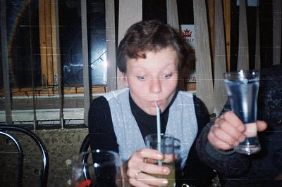 Vintage indoor shot of a boy drinking from a straw in a glass, surrounded by empty bottles and a blurred wooden railing. Mid-...