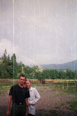 Vintage photo of two individuals standing in a rural, rain-soaked landscape. The man wears a dark shirt and pants, the woman ...