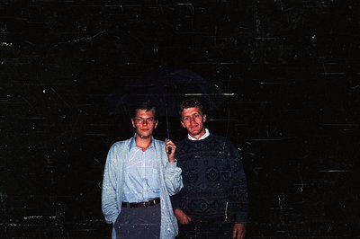 Two men pose indoors against a dark, textured wall. The man on the left wears a light blue button-down shirt, glasses, and a ...
