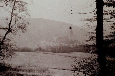 Vintage sepia-toned photograph of a mist-shrouded hillside with faint ruins or industrial structures partially obscured by fo...