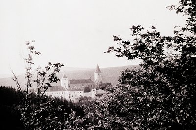 Mid-20th century black-and-white shot of a fortified castle nestled in a valley, partially obscured by foliage. Distinctive c...