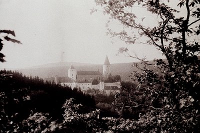 Vintage sepia-toned photograph of a hilltop castle complex with a central tower and surrounding walls, framed by foliage. Lik...