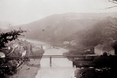Vintage black-and-white aerial view of a steel truss bridge spanning a winding river, with a grand, multi-story building near...
