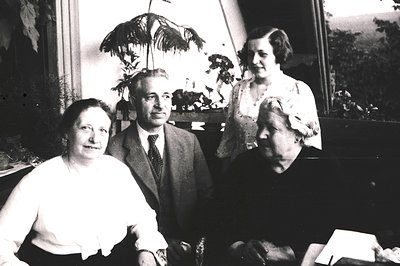 Vintage black-and-white portrait of four individuals in a formal indoor setting, likely mid-20th century. The man in the cent...