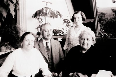 Four individuals pose indoors, likely mid-20th century (1940s–1950s). A man in a striped tie stands between two women in form...