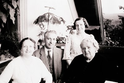 Four-generation family portrait in early 20th-century indoor setting, likely 1920s–1930s. Three women and one man pose near l...