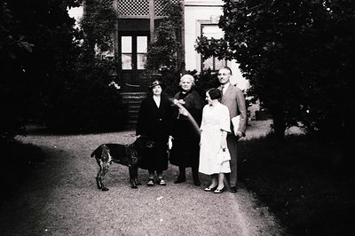 Family portrait in early 20th-century European estate setting. Four adults and a child pose outdoors near a grand house with ...