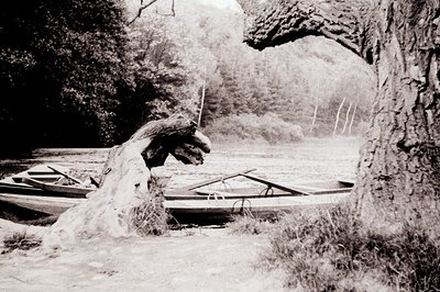 Black-and-white photograph of two wooden boats resting on a rocky riverbank beneath a natural rock arch. Overgrown vegetation...