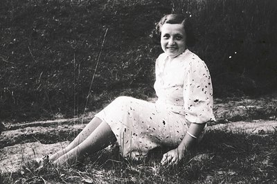 Young woman in 1940s-50s dress poses outdoors on grassy terrain, surrounded by sparse vegetation. Her floral-patterned blouse...