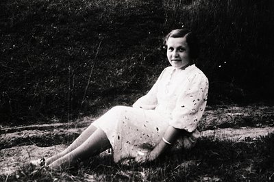 Young girl in a floral dress poses outdoors, seated on a rough-hewn wooden bench against a textured stone wall. Mid-20th cent...