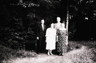 Four individuals pose outdoors in a wooded area, likely mid-20th century (1950s–1960s). Men wear suits, women in formal dress...