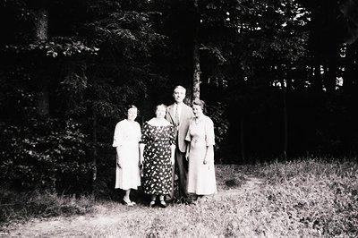 Four individuals pose outdoors in a wooded area, likely mid-20th century (1940s–1960s). The man in the center wears a suit wi...