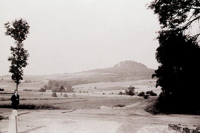 Vintage sepia-toned landscape featuring a lone figure in traditional attire standing near a rural roadside fence. Rolling fie...