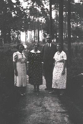Four individuals pose outdoors in a wooded area, likely mid-20th century (1940s–1950s). The man wears a suit with a tie, flan...