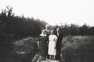 Four individuals pose outdoors on a gravel path, surrounded by dense forest. The woman on the left wears a polka-dot dress; t...