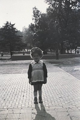 Black-and-white portrait of a young girl in a wide-brimmed hat and patterned dress, standing on a paved plaza surrounded by t...