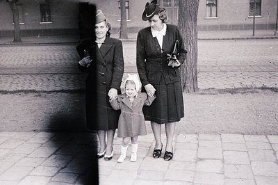 Three-generation family portrait in mid-20th century urban setting. Woman in 1940s-style hat and coat holds a young girl in a...