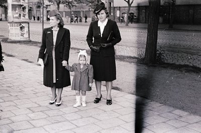 Three women in mid-20th-century attire—wide-brim hats, knee-length coats, and knee-high socks—walking on a paved urban street...