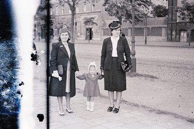 Vintage black-and-white street scene featuring two women in 1940s-era suits and hats, holding hands with a young girl in a dr...