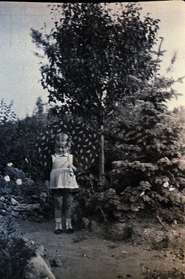 Vintage black-and-white photo of a young girl in a simple dress and knee-high socks standing beside a tree on a rocky path, m...