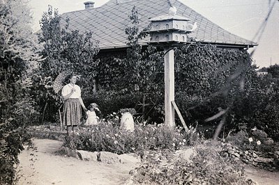 Vintage sepia-toned garden scene featuring a woman and child beside a wooden birdhouse perched on a stone pedestal. The house...