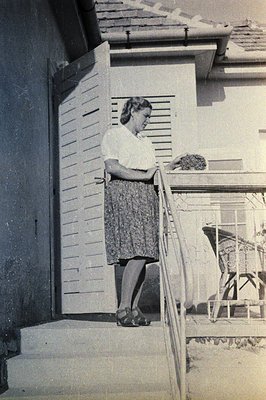 Mid-century woman in a skirt and blouse stands on brick steps beside a wrought-iron railing, holding a book. Residential exte...