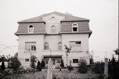 Three-story Art Nouveau-inspired villa with mansard roof, symmetrical facade, and arched windows. Overgrown shrubs and barbed...