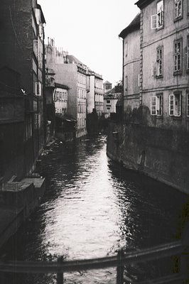 Narrow urban canal flanked by multi-story residential buildings with shuttered windows, likely mid-20th century. Overcast ski...