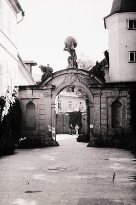 Baroque-style stone gateway with sculpted urn atop archway, flanked by ornate carvings. Two figures walking through courtyard...