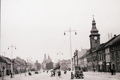 Black-and-white street scene featuring early 20th-century European architecture. Prominent clock tower with ornate spire domi...