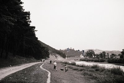 Black-and-white rural scene: winding dirt road flanked by dense forest on left, leading to a riverbank with three figures—two...