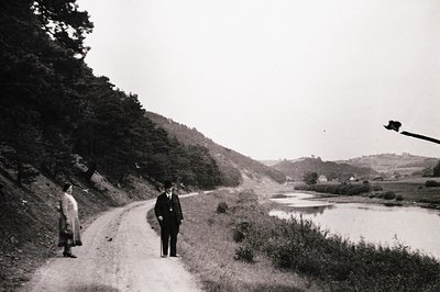 Two figures walk a rural dirt road beside a shallow stream, framed by dense greenery and rolling hills. The man in a suit and...