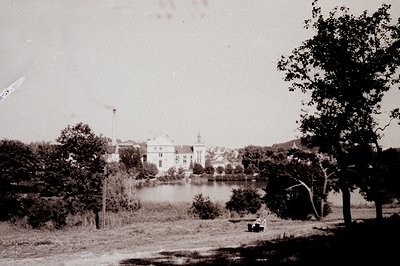 An expansive view of a grand, ornate building reflecting in a calm lake. Lush greenery frames the water's edge; a small bench...