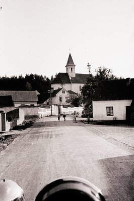 Quaint village scene: a narrow road leads to a church with a steeple, framed by traditional houses and a low fence. Several f...