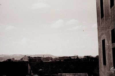 A rooftop cityscape with a prominent building facade framing the view. Distant hills and a visible monument characterize the ...